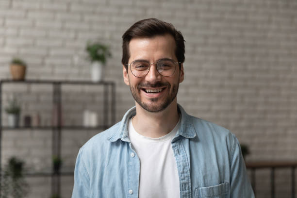 istockphoto-1300701503-612×612 Headshot portrait of happy young man looking at camera standing indoors at home or modern office. Profile picture of smiling millennial male making online call by video link posing for digital webcam