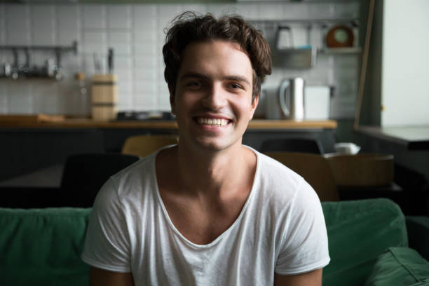 istockphoto-974371470-612×612 Smiling millennial man head shot portrait, happy handsome young blogger looking at camera sitting on kitchen sofa making video call or shooting blog, cheerful casual friendly guy posing at home